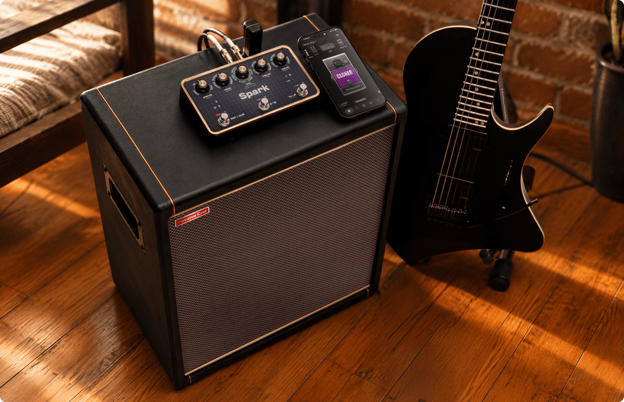 Spark PEDAL sitting on top of a Spark CAB 2 on a wooden floor, with a phone running the Spark app and a guitar beside it